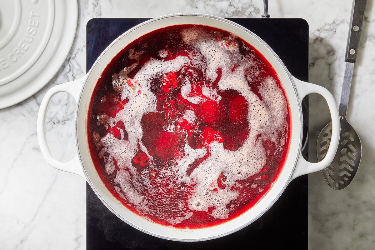 A white pot filled with bubbling red liquid, possibly simmering fruit or berry jam, sits on a stovetop with a slotted spoon nearby.