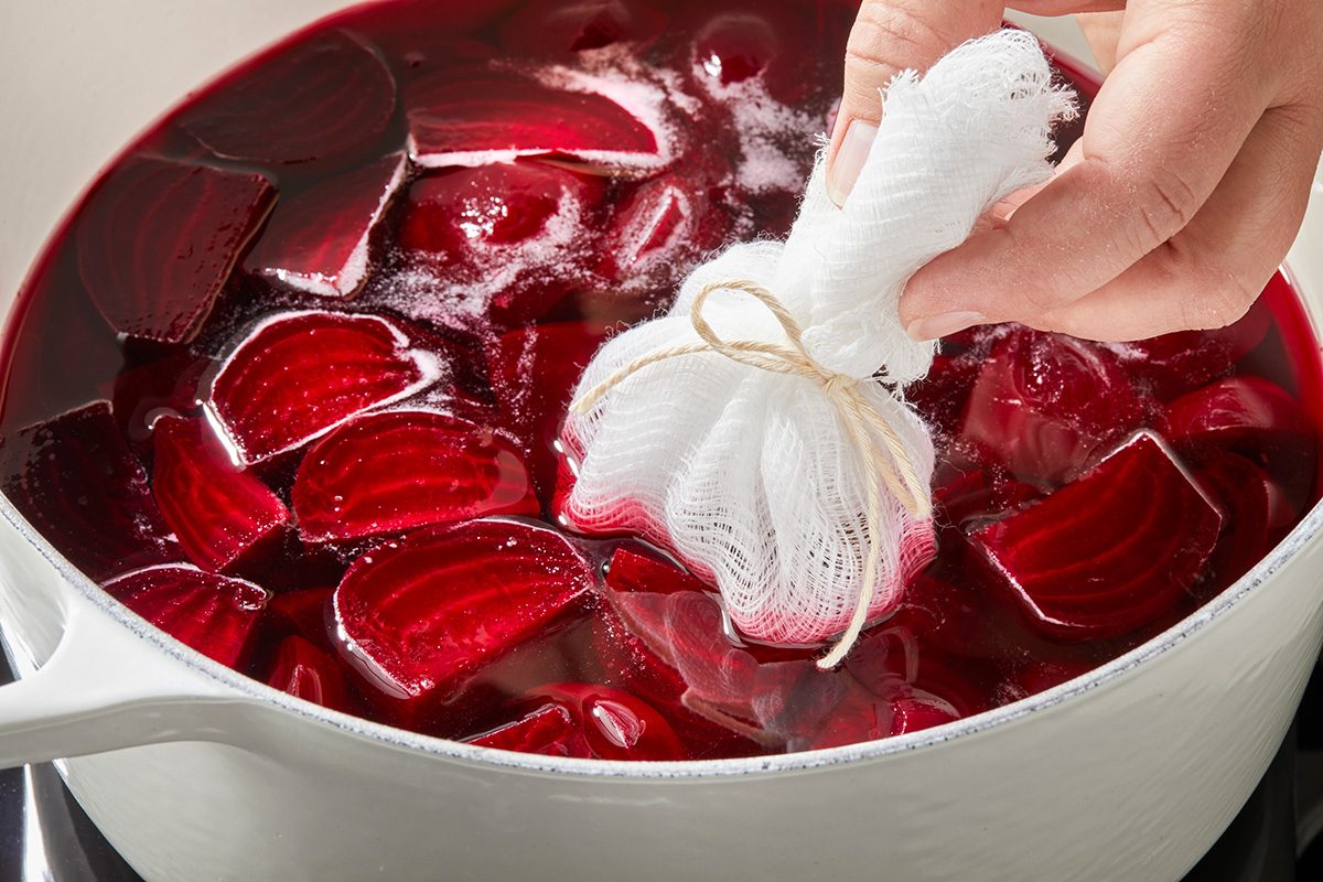 A hand lowers a bundle of spices wrapped in cheesecloth into a pot filled with chopped red beets and liquid.