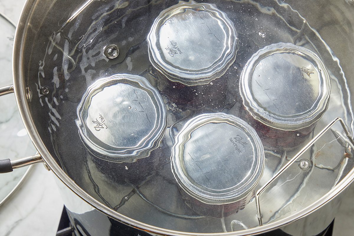 Four glass jars with metal lids are being processed in a large pot of boiling water for canning, viewed from above. A metal rack is partially visible inside the pot.