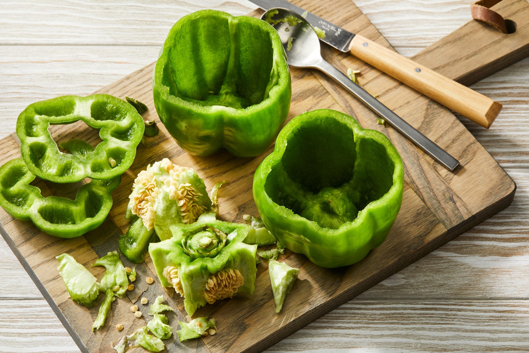 Overhead shot of removing seeds from peppers on a wooden cutting board; with a spoona nd knife; all set on a cream colored wooden surface;
