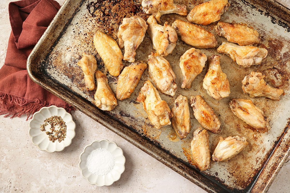 overhead shot of a baking sheet filled with golden brown baked chicken wings sits on a light surface next to a brown napkin, Two small dishes hold salt and a pepper blend