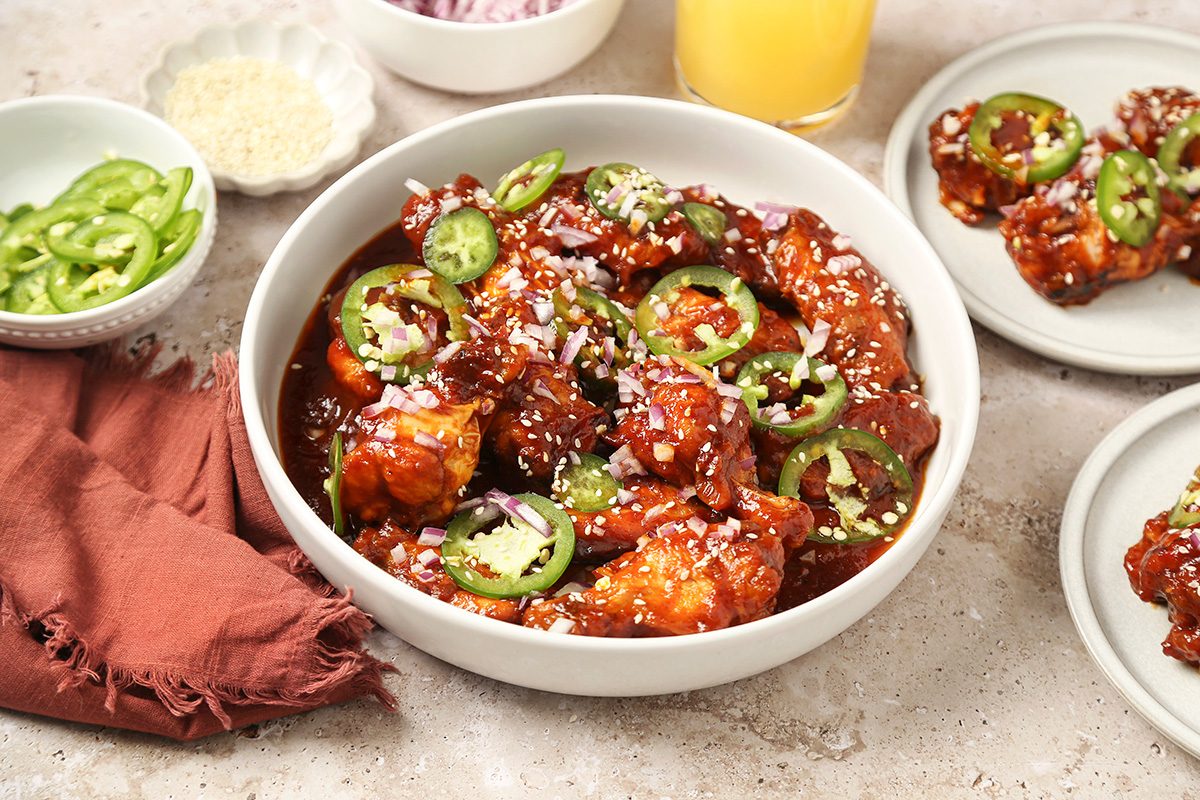 3/4th shot of a bowl of spicy glazed chicken pieces topped with sliced green chilies, chopped onions, and sesame seeds, surrounded by small bowls of sliced chilies, sesame seeds, onions, a drink, and a rust colored napkin