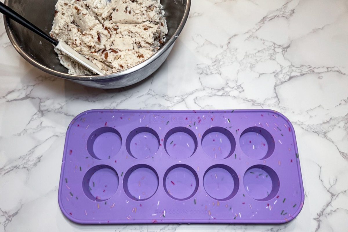 A purple silicone mold with ten round cavities sits on a marble counter next to a metal mixing bowl filled with a creamy, speckled mixture and a spatula.