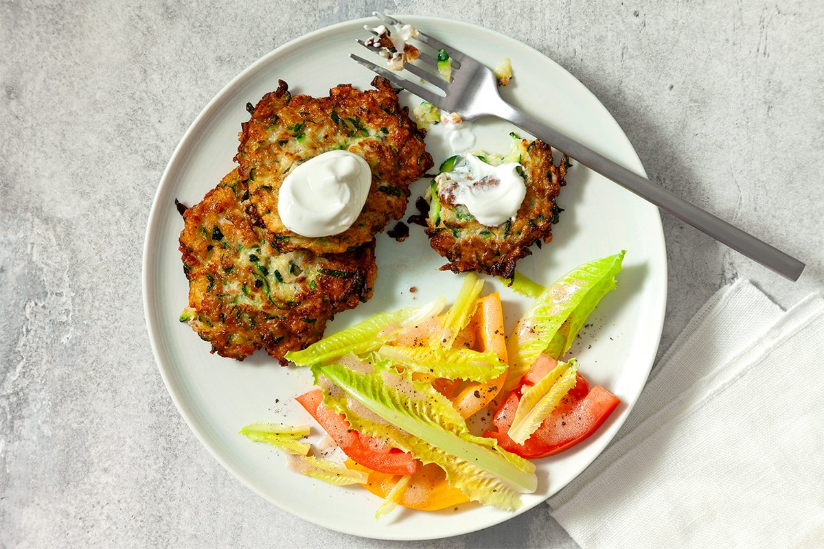 A white plate with three zucchini fritters topped with sour cream, served alongside a fresh salad of lettuce, tomato, and yellow bell pepper. A fork rests on the plate with a bite of fritter on it.