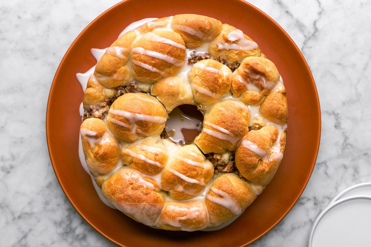 overhead shot of a round pull apart apple bread ring with golden brown rolls, drizzled with white icing, sits on an orange plate atop a marble surface