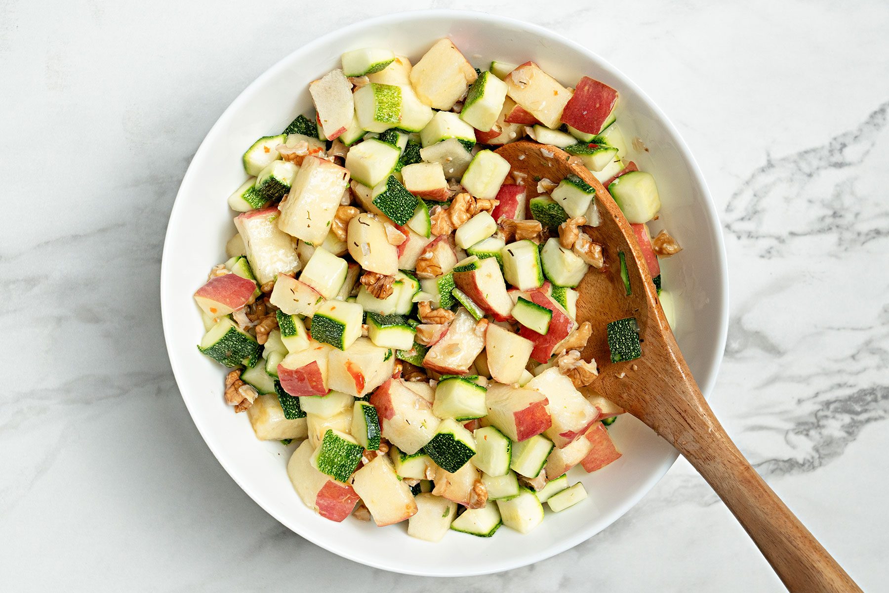Apples, zucchini and walnuts combined with italian seasoning in a large bowl with a wooden ladle.