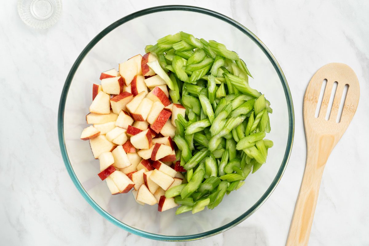 Overhead shot of a glass bowl with celery and apples tossed in lemon juice; a flat wooden spoon nearby on a marble surface;