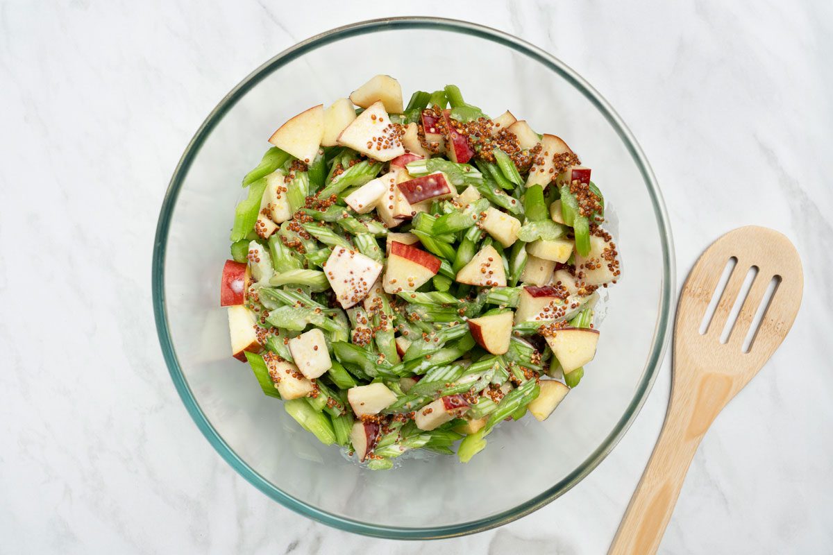 Overhead shot of dressing being poured over the celery mixture; flat wooden spoon nearby on a marble surface;