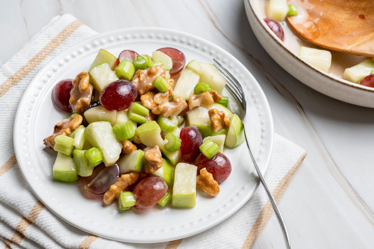Top view shot of Apple Grape Salad in a large bowl with wooden serving spoon; served on a plate with a fork over a napkin nearby; all set on a marble surface;
