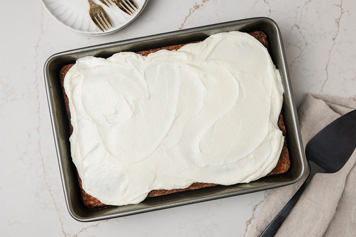 A rectangular cake in a baking pan topped with smooth white frosting, placed on a marble surface. A plate with forks and a black serving spatula are nearby.