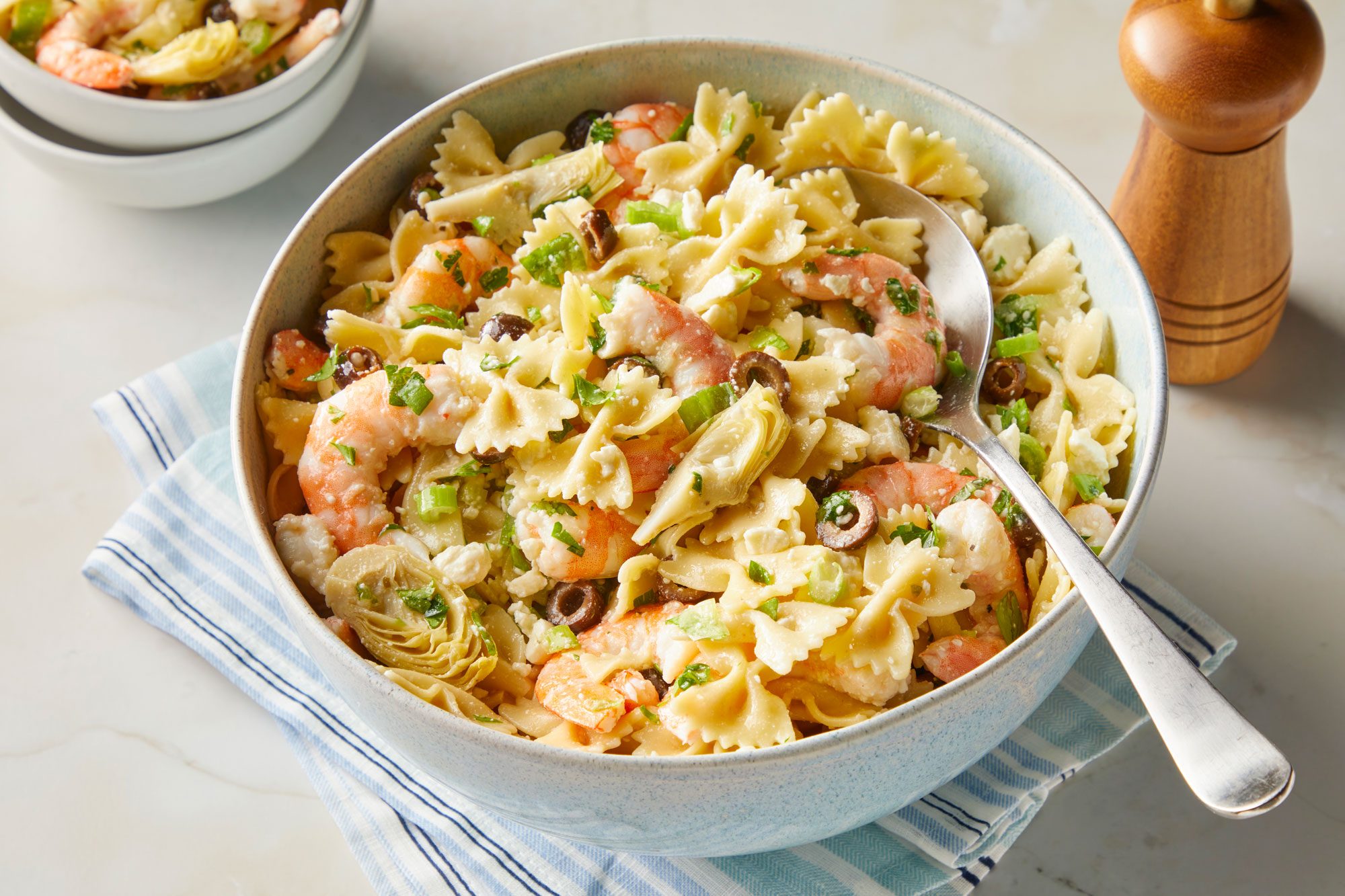 3/4 angle view shot of Artichoke Shrimp Pasta Salad in a large bowl with a serving spoon placed over a napkin; and a small serving bowl nearby; all set on a light marble surface;