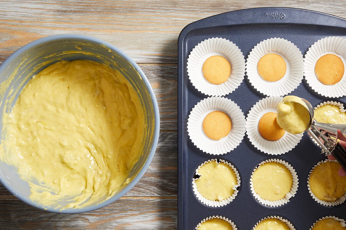 A bowl of yellow batter sits next to a muffin tin lined with paper cups, each holding a vanilla wafer. A spoon is used to scoop batter into the cups.