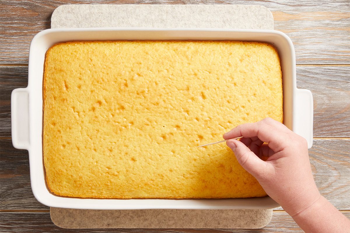 A hand is inserting a toothpick into a rectangular yellow cake in a white baking dish, checking if it is fully baked. The dish is on a wooden surface.