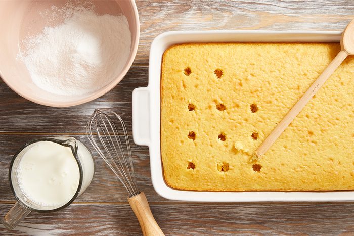 A rectangular baked cake in a white dish with holes poked in the top, next to a wooden spoon, a whisk, a bowl of powdered sugar, and a glass measuring cup of milk on a wooden surface.