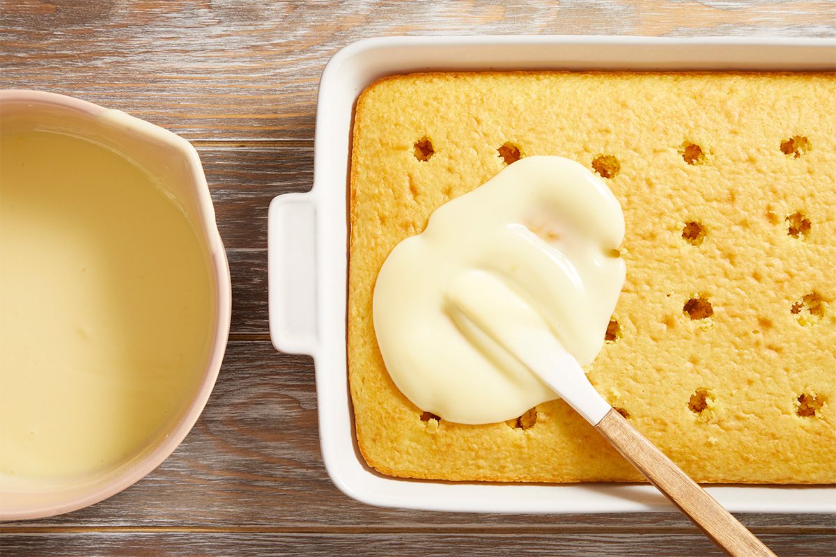 A rectangular yellow cake in a white baking dish has holes poked in it and is being topped with creamy sauce using a spatula. A bowl of extra sauce sits beside the dish on a wooden surface.