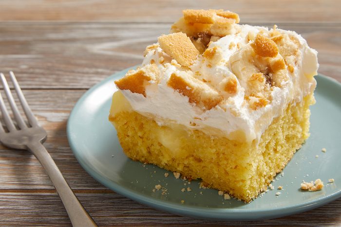 A slice of yellow cake topped with whipped cream and crumbled cookies sits on a green plate, with a fork beside it on a wooden table.