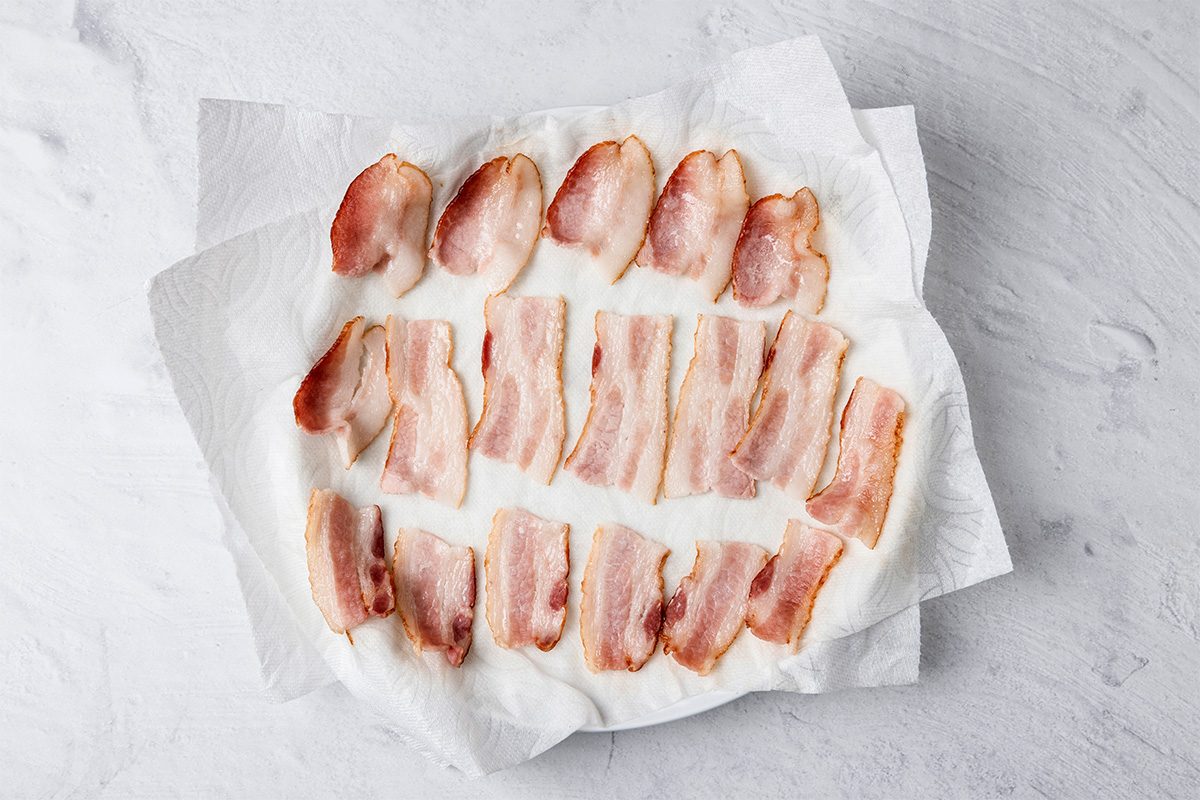 Cooked bacon strips arranged on a plate lined with white paper towels, set on a light gray surface. The paper towels are absorbing excess grease from the bacon.