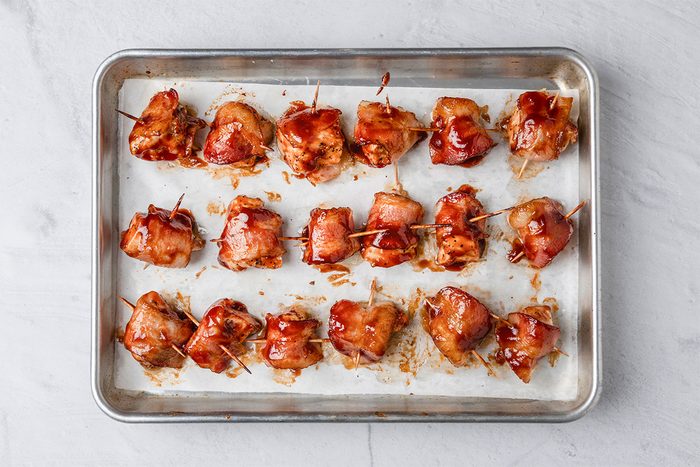 A baking sheet lined with parchment paper holds several pieces of bacon-wrapped chicken bites, each secured with a toothpick and coated in a glossy sauce.