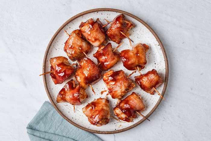 A plate of bacon-wrapped appetizers secured with toothpicks and glazed with a reddish sauce, arranged neatly on a white plate with a brown rim, next to a folded blue napkin on a light surface.
