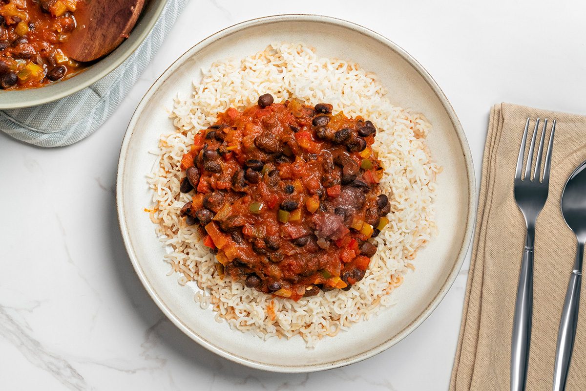 A plate of white rice topped with a hearty black bean and vegetable stew, served next to a fork, knife, and napkin on a white surface.