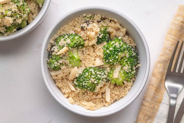 A white bowl filled with couscous, broccoli florets, and slivered almonds sits on a white surface next to a fork and a beige-striped napkin.