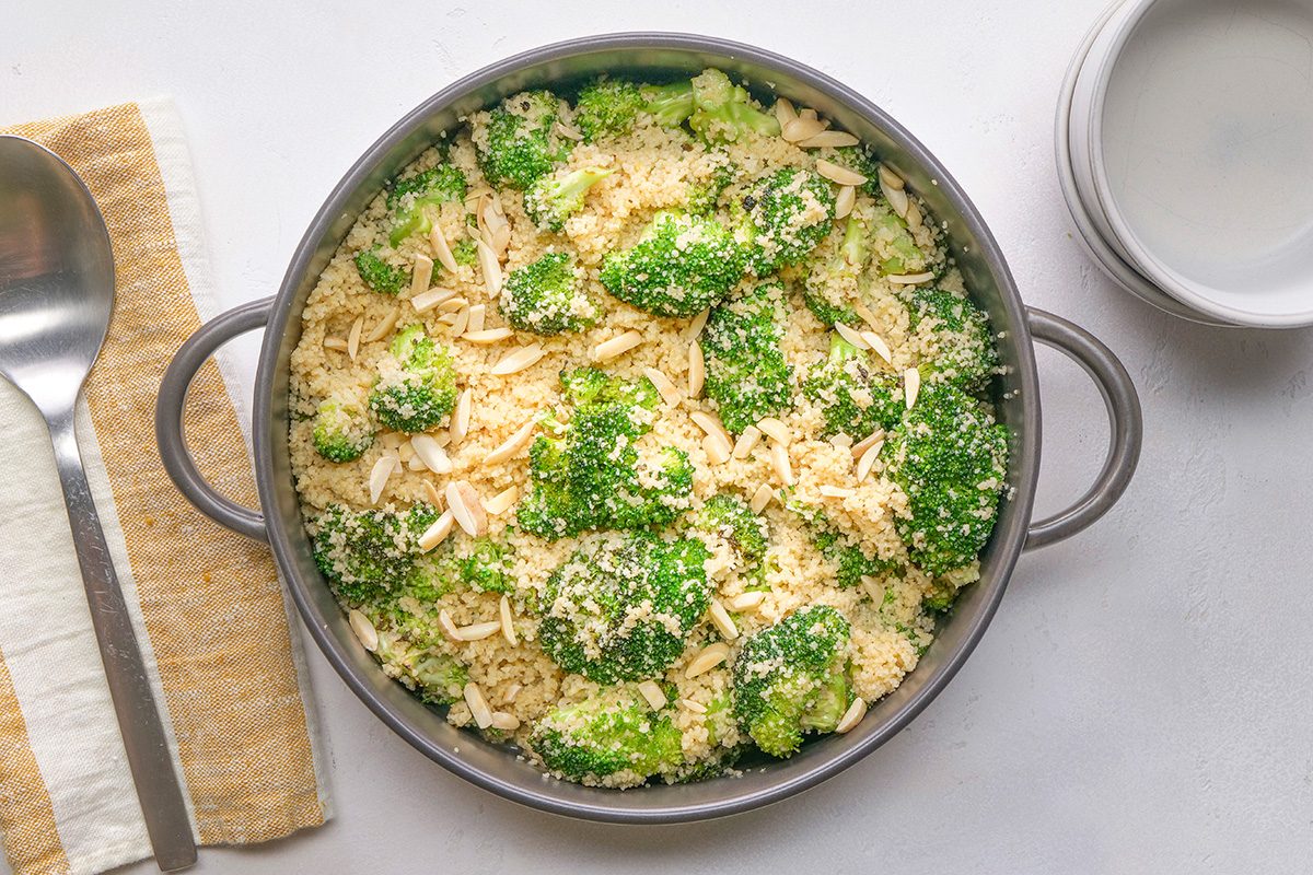 A round baking dish filled with broccoli florets topped with breadcrumbs and shredded cheese sits on a light surface next to a spoon, a napkin, and a small empty bowl.