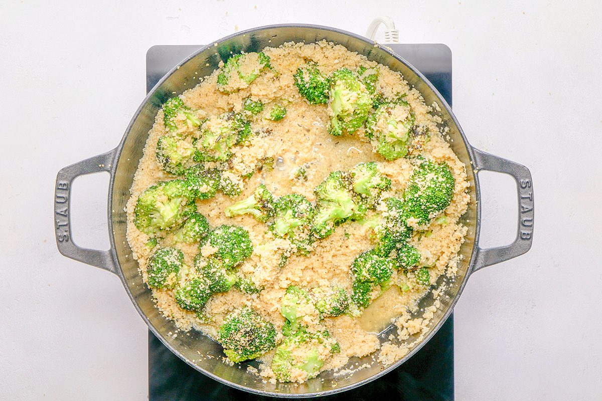 A large skillet filled with broccoli florets and breadcrumbs on top, cooking on a stovetop with a white background.