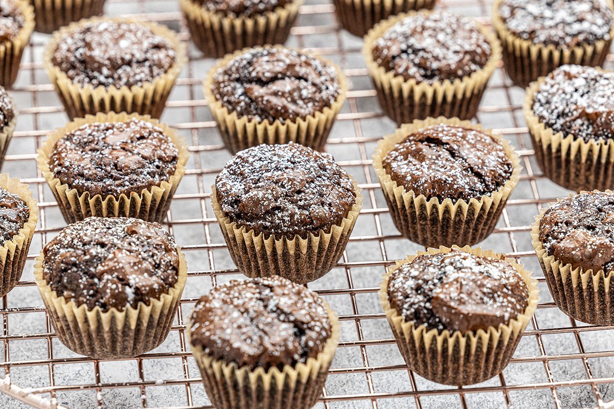 Rows of chocolate muffins in paper liners are cooling on a wire rack, topped with a dusting of powdered sugar.