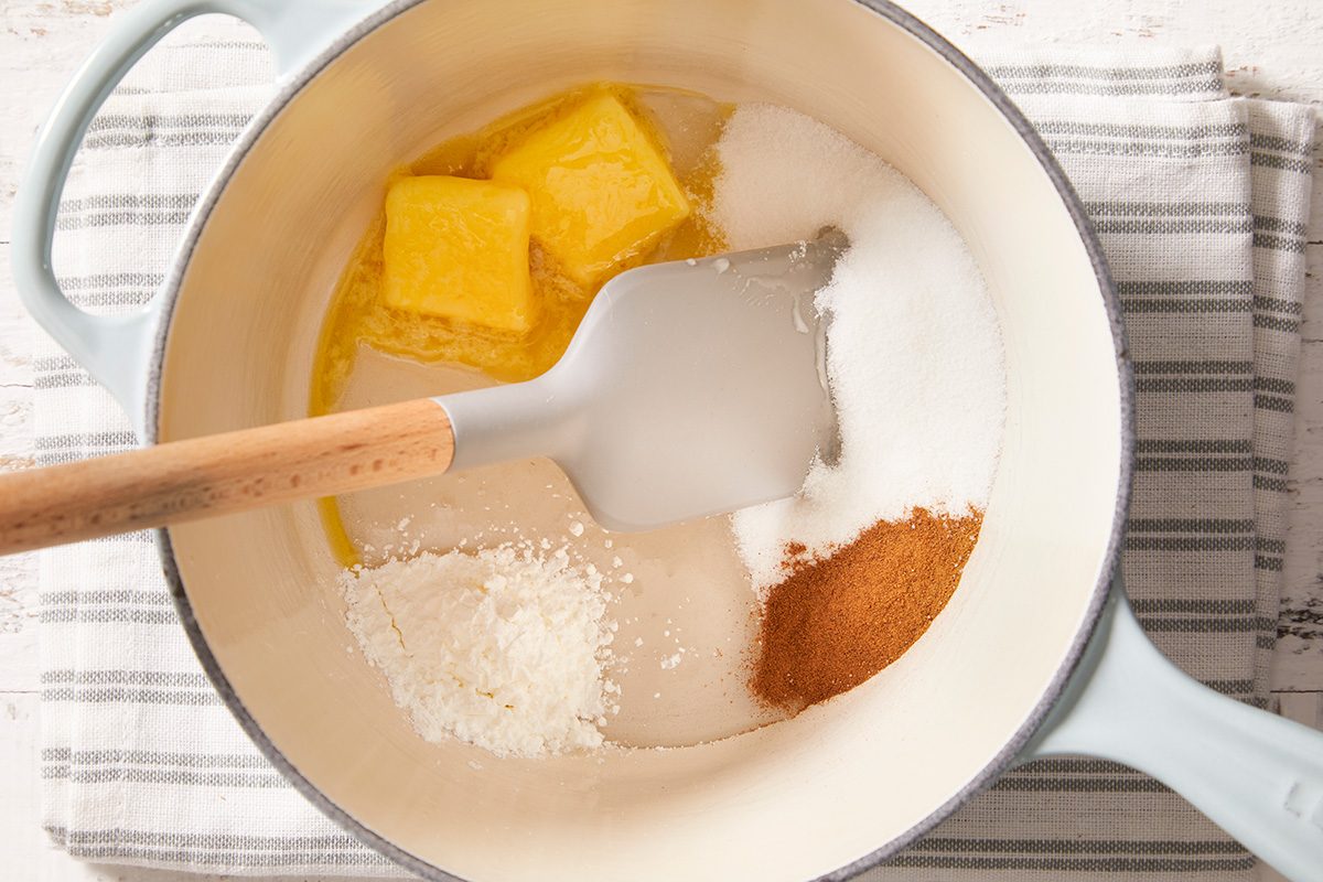 A saucepan with butter, sugar, cinnamon, and other baking ingredients being mixed with a spatula, set on a striped cloth.