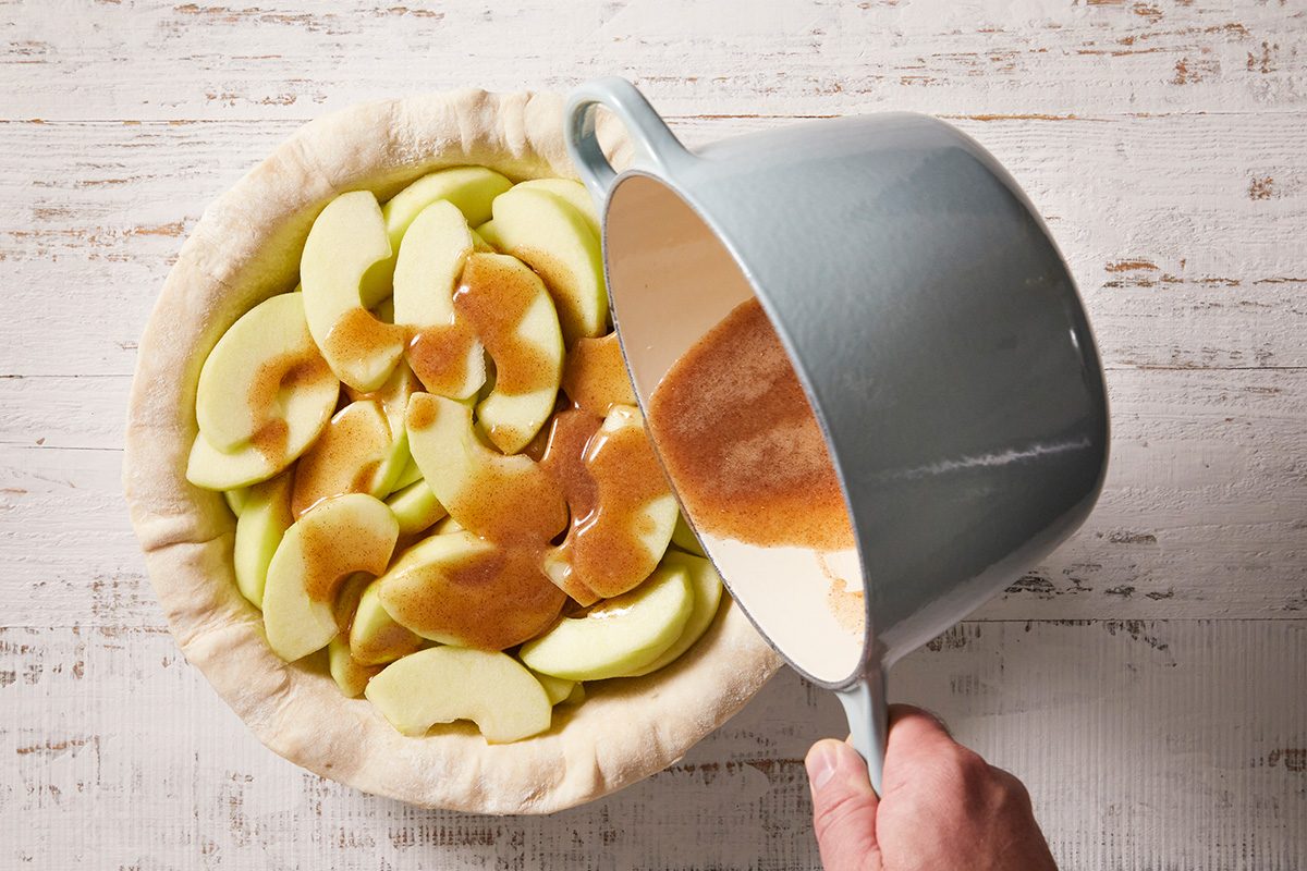 A hand pours caramel-colored sauce from a saucepan over sliced green apples arranged in a pie crust, preparing an apple pie on a rustic white wooden surface.
