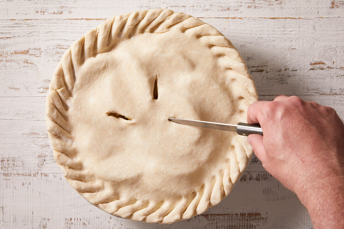 A hand holds a knife, cutting slits into the top crust of an unbaked pie with crimped edges on a white wooden surface.