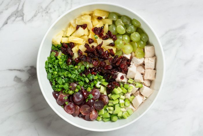 Overhead shot of a large bowl combine the chicken, pineapple, celery, onions, grapes and cranberries; on a marble surface