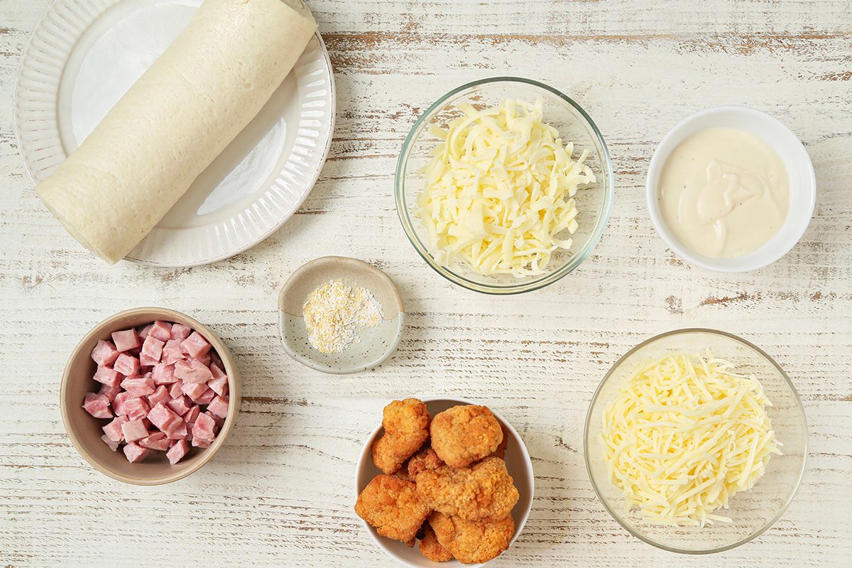 A flatlay of ingredients on a white wooden surface, including a tortilla wrap, two bowls of shredded cheese, a bowl of diced ham, breaded chicken pieces, grated Parmesan, and a bowl of white sauce.