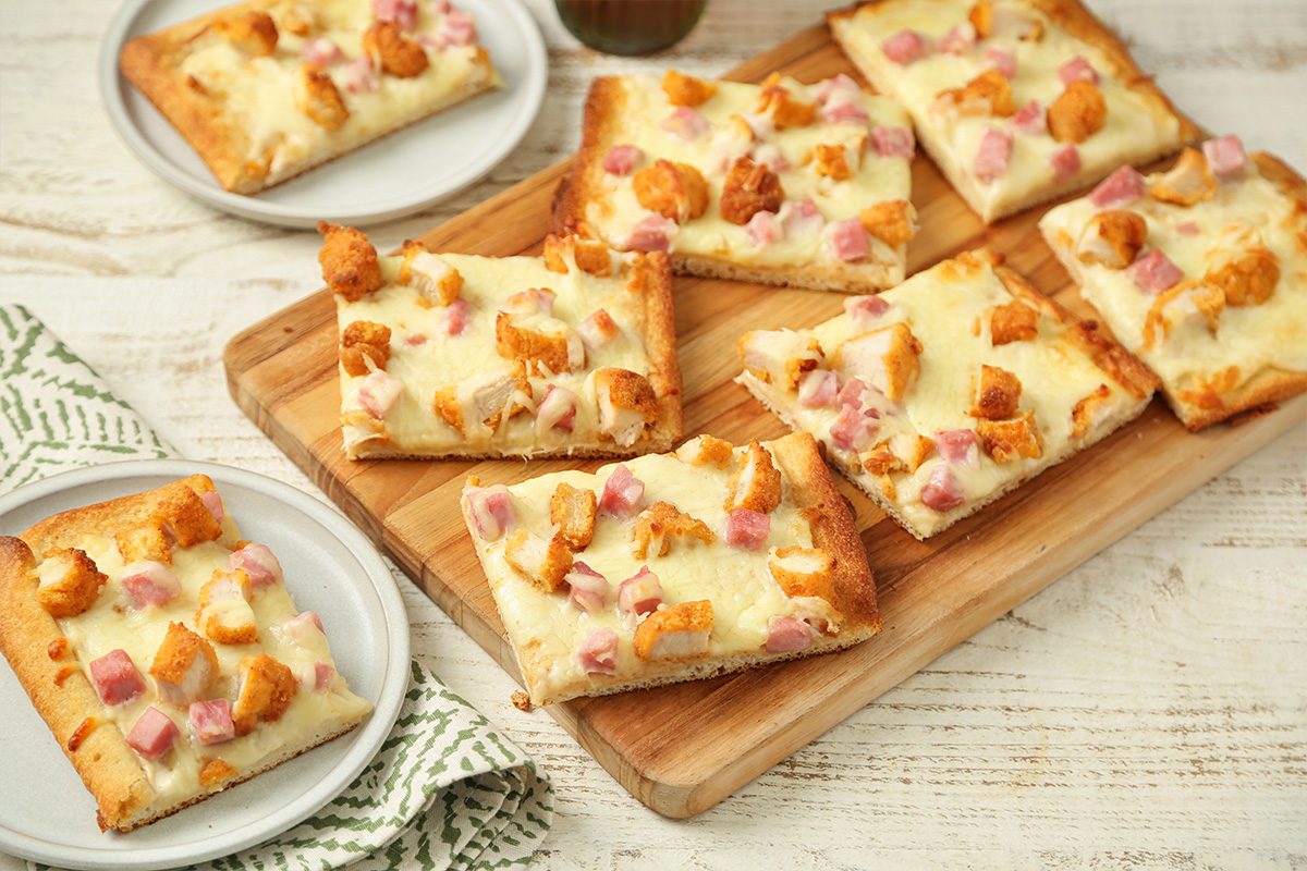 Rectangular slices of flatbread topped with melted cheese, diced ham, and chicken pieces are arranged on a wooden board. Two additional pieces are served on small round plates.