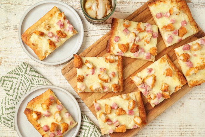 Rectangular slices of baked pizza with cheese, diced ham, and chicken are arranged on a wooden board, with two slices on separate white plates nearby. A patterned napkin and a glass jar are also visible on the table.