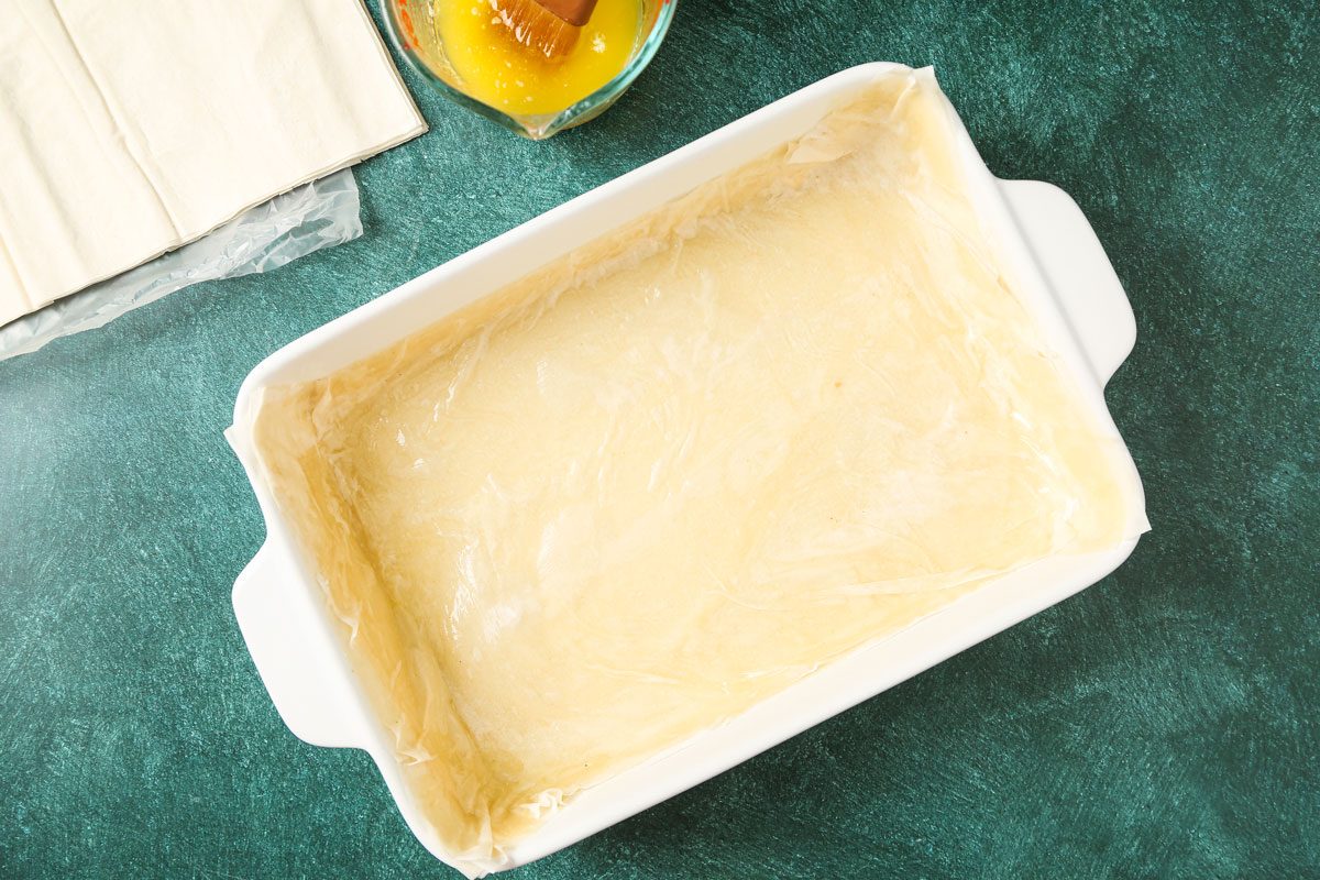 A white rectangular baking dish with phyllo dough layers is on a green countertop; extra phyllo sheets and a bowl of yellow fruit in syrup are beside it. Preparation for a dessert is in progress