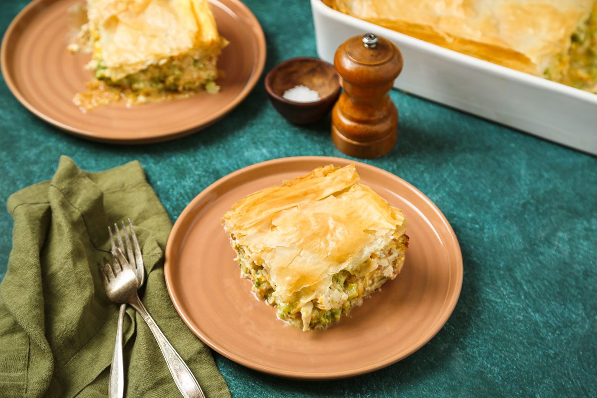 A slice of savory pie rests on a tan plate; golden flaky crust glistens; background features utensils; salt; pepper grinder; another plate; and the remaining pie in a white baking dish. Cozy dining scene