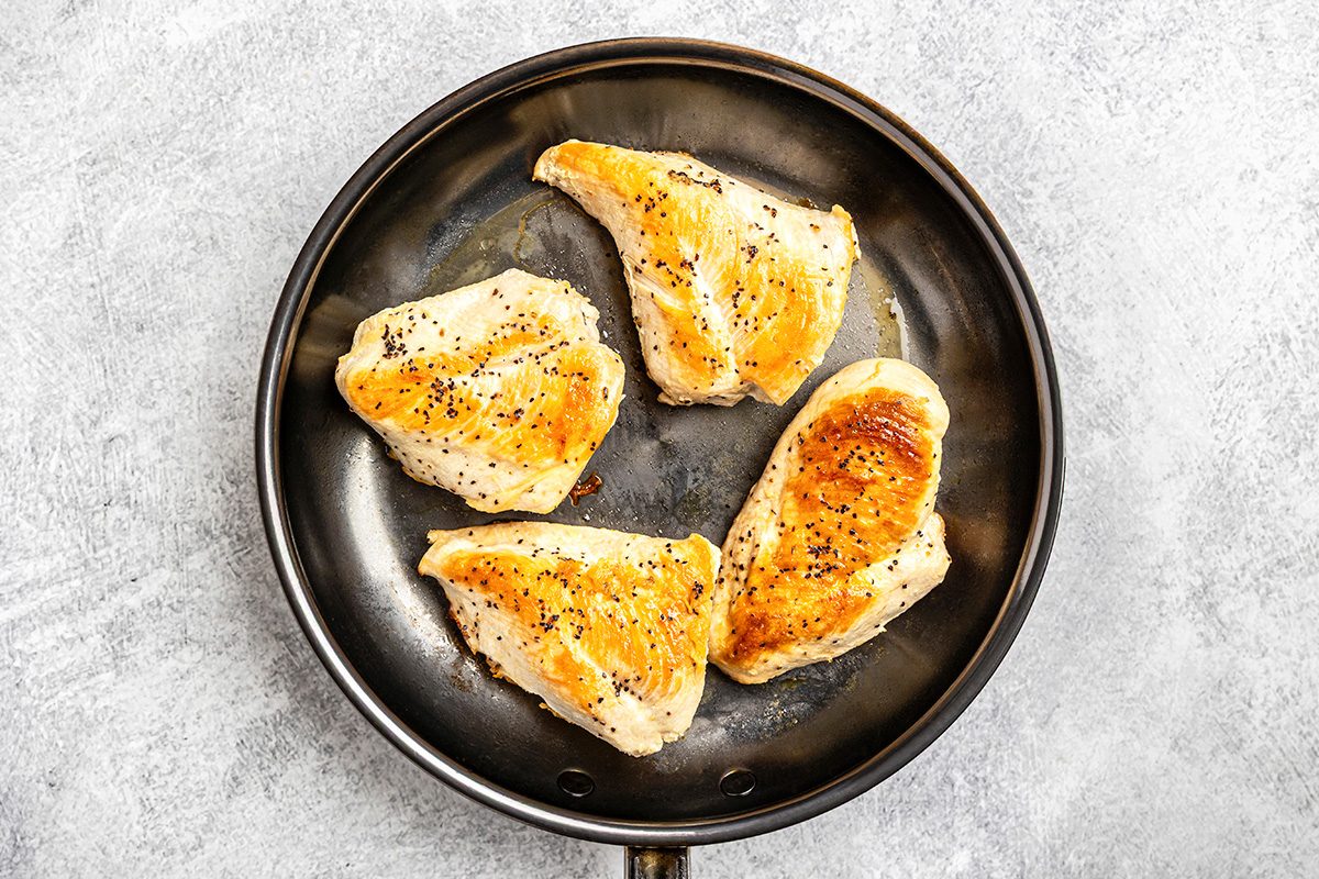 Four seasoned, golden-brown chicken breasts cooking in a black frying pan, set on a light gray textured surface.