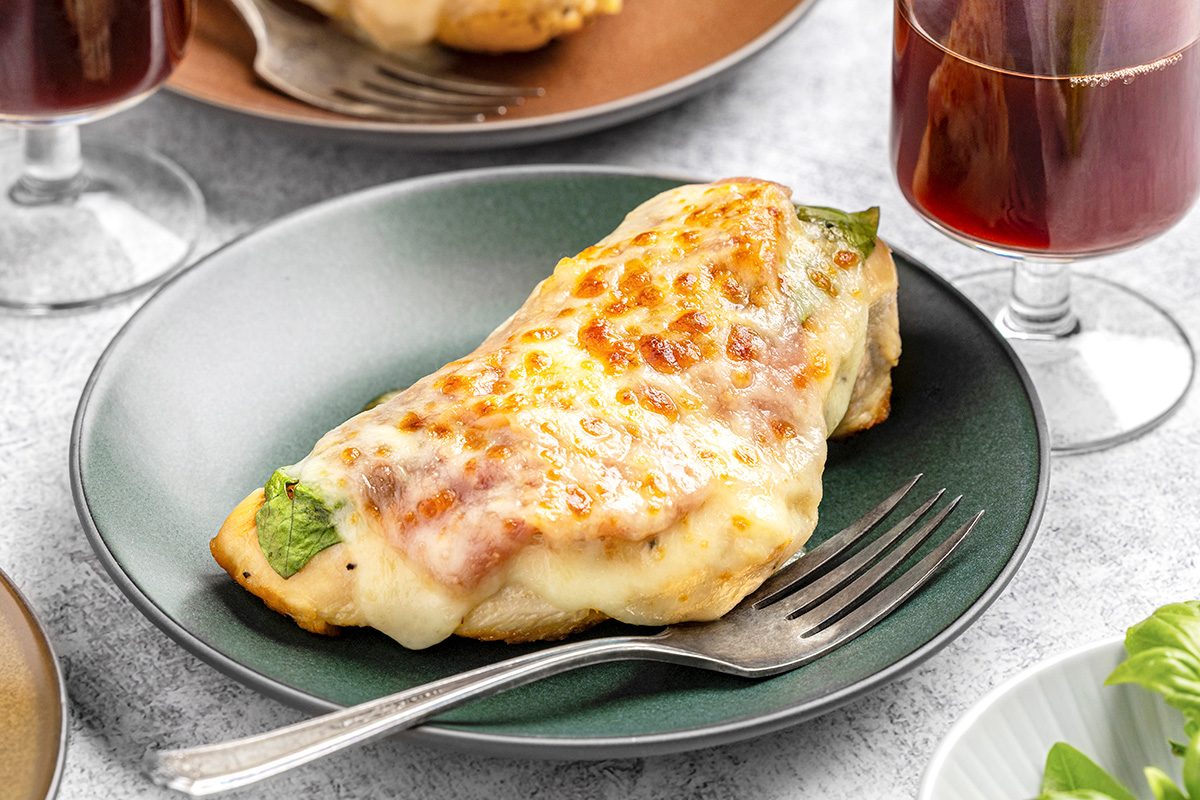 A plate with a piece of baked chicken breast covered in melted cheese and herbs, served with a fork. Two glasses of red wine are nearby on the table.