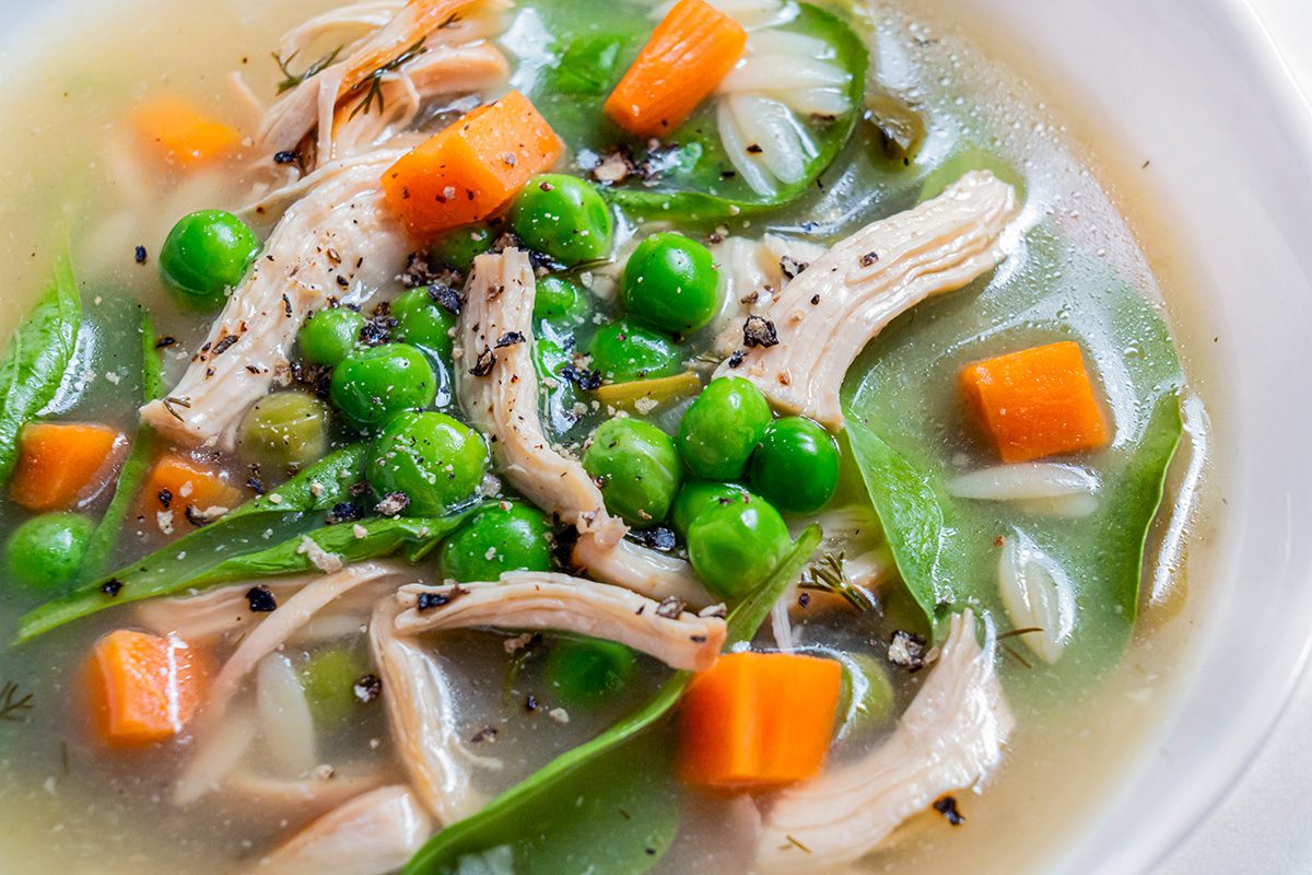 A close-up of a bowl of chicken soup with shredded chicken, green peas, carrot cubes, snap peas, onion slices, and black pepper in a clear broth.