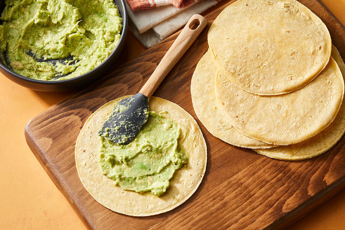 A wooden board with several corn tortillas; one tortilla is being spread with guacamole using a spatula. A bowl of guacamole and a stack of tortillas are nearby.