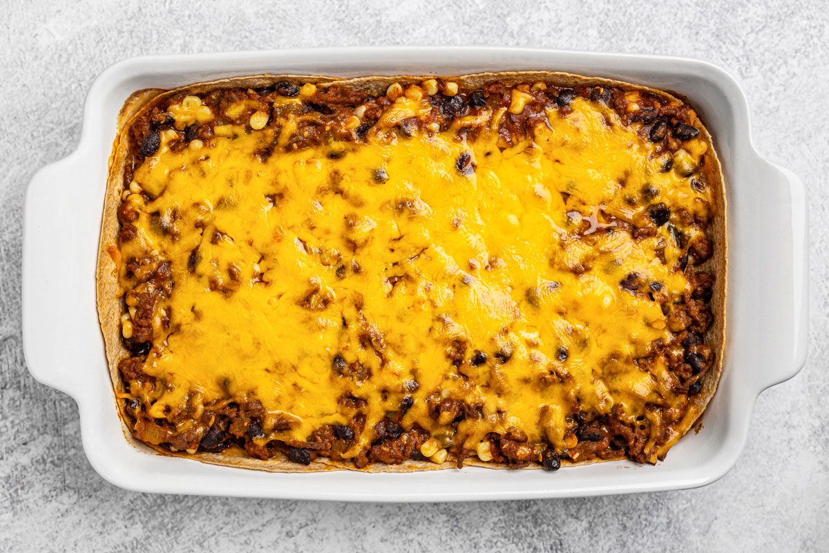 overhead shot of a white rectangular baking dish filled with a baked casserole topped with melted cheddar cheese, ground meat, and beans, set on a light gray textured surface