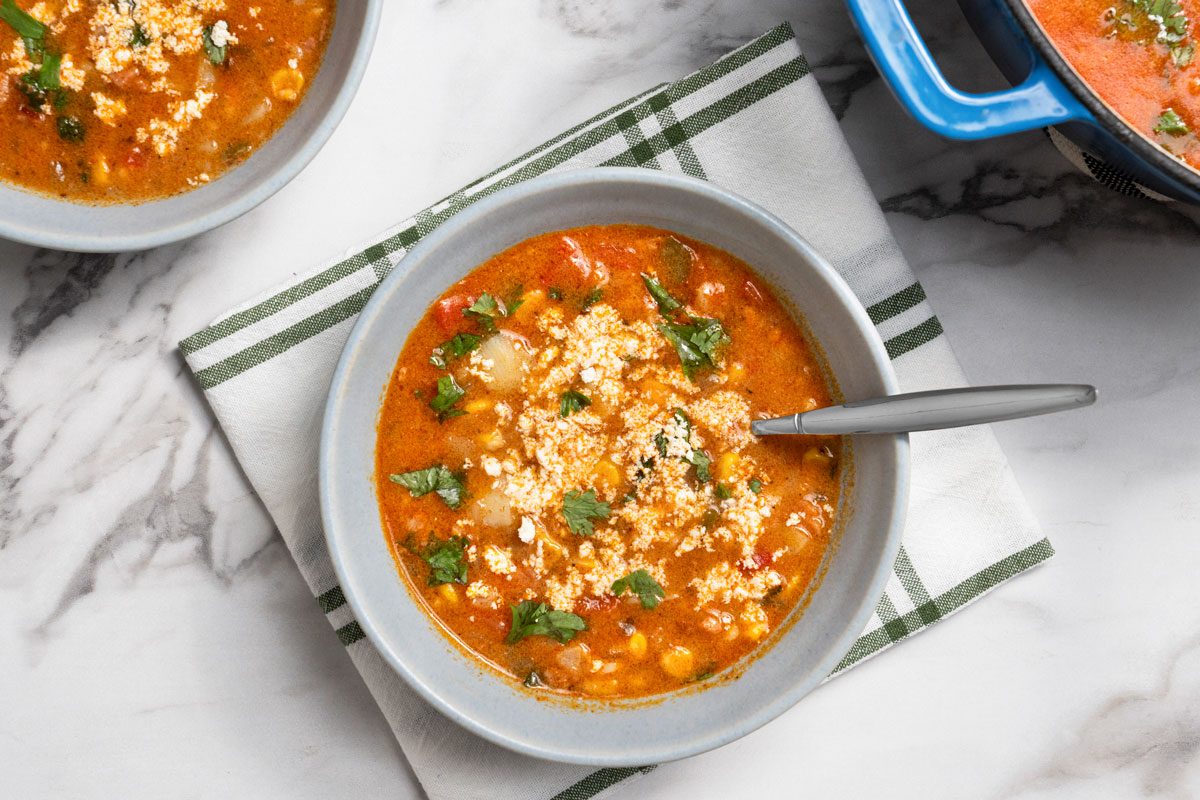 Overhead shot of Mexican Chorizo and Corn Soup served in two light gray bowls; The bowl is placed on a white cloth napkin with green stripes; A spoon rests inside the bowl; all set on a marble surface;