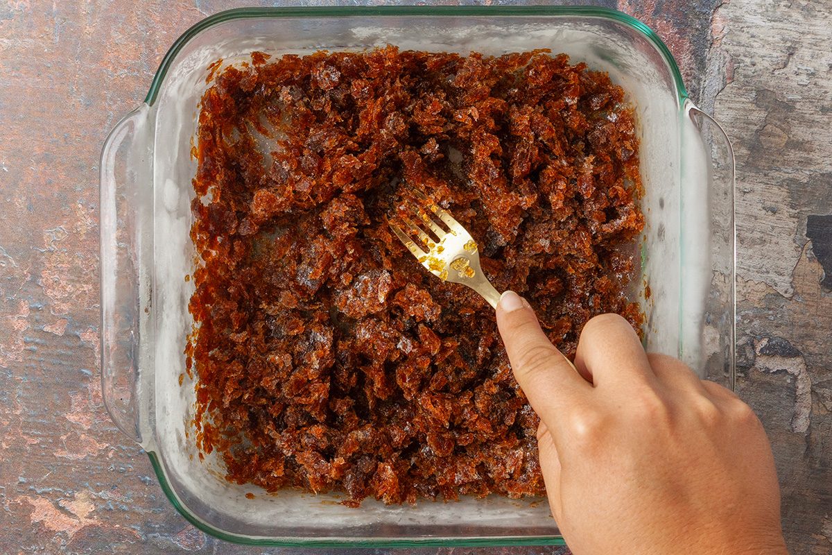 overhead shot of a hand uses a fork to mash a thick, dark brown mixture in a glass baking dish on a rustic surface
