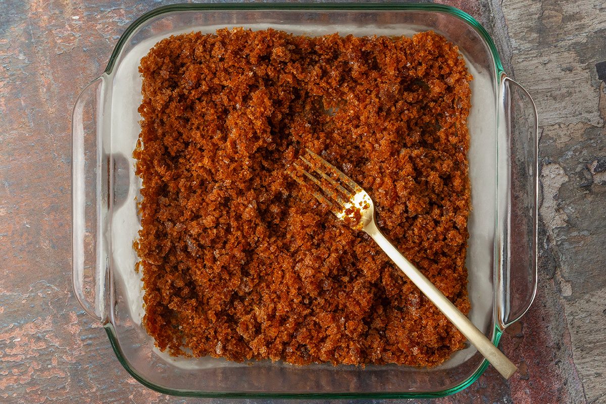 overhead shot of a glass baking dish filled with a coarse, brown, granulated mixture; the dish sits on a rustic surface with shades of brown, gray, and blue creating a textured background