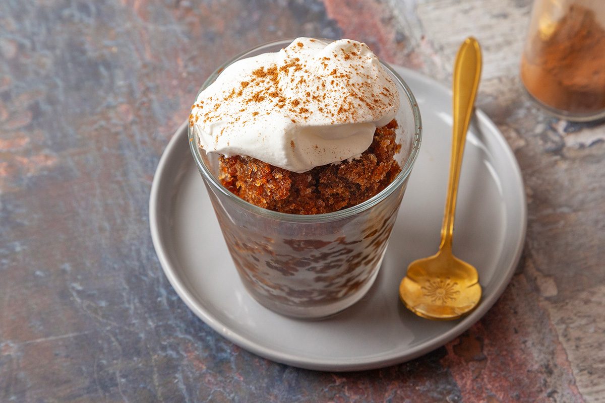 3/4th shot of a glass filled with a layered dessert of whipped cream and what appears to be spiced cake crumbs, topped with more cream and a sprinkle of cinnamon, sits on a small gray plate with a gold spoon beside it