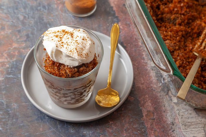 3/4th shot of a glass filled with layered dessert topped with whipped cream and sprinkled cinnamon sits on a plate with a gold spoon; A baking dish with more dessert and a jar of cinnamon are nearby on a textured surface