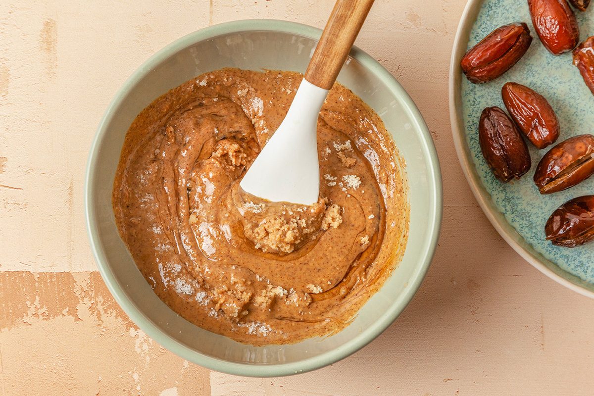 Overhead shot of slice dates lengthwise to create a small pouch; In a small bowl stir together almond flour, almond butter, maple syrup, vanilla extract and salt until a soft dough forms; spatula; all set on a light orange background;