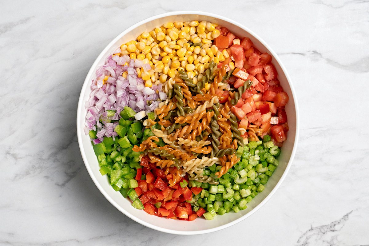 A white bowl filled with spiral pasta, diced tomatoes, cucumbers, red onions, bell peppers, and corn, arranged in colorful sections on a marble surface.