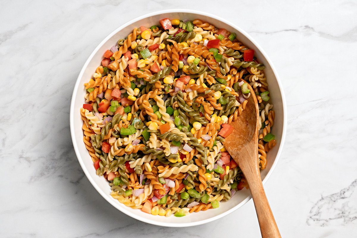 A bowl of colorful pasta salad with chopped vegetables, including tomatoes and green peppers, is shown on a marble surface. A wooden spoon rests inside the bowl.